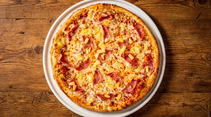 A whole Hawaiian pizza with ham and pineapple on a white plate, on a wooden table.